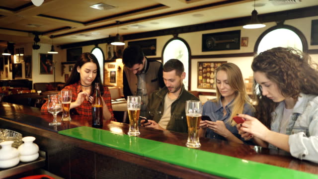 Young-people-are-checking-smartphones-while-sitting-at-bar-counter-together.-They-are-showing-each-other-screens,-laughing-and-talking.-Modern-technologies-communication-concept.
