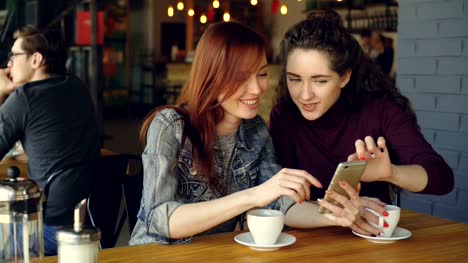 Pretty-young-girl-is-using-smartphone-and-showing-interesting-photos-to-her-female-friend-the-discussing-them-while-drinking-coffee-in-nice-modern-cafe.