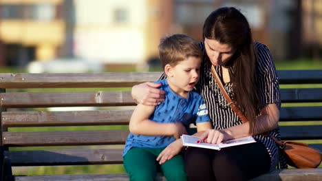 Young-mother-and-son-sitting-on-a-bench-in-a-park-and-reading-a-book