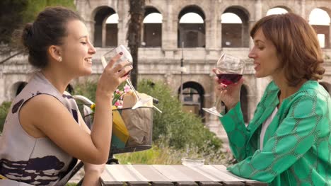 Two-happy-young-woman-tourists-sitting-at-the-table-of-a-bar-restaurant-in-front-of-the-Colosseum-in-Rome-drink-and-toast-with-a-glass-of-italian-red-wine.-Stylish-colorful-dress-on-a-summer-day-at-sunset