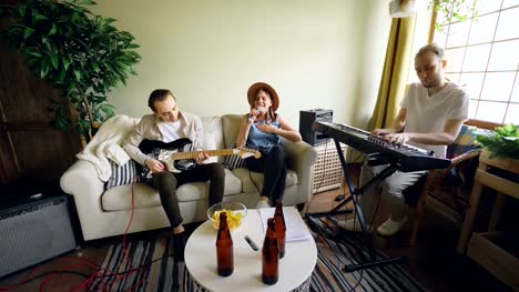 Musikband-zu-Hause-üben,-Frau-singt-und-Männer-spielen-Musikinstrumente-Gitarre-und-Keyboard.-Flaschen-Bier-und-Snacks-auf-modernen-Tisch-sind-sichtbar.