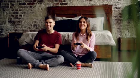 Nettes-junges-paar-Freund-und-Freundin-spielen-Videospiel-Holding-Joysticks-auf-dem-Boden-des-Schlafzimmers-zu-Hause-sitzen.-Emotionale-Liebhaber-erfreuen-sich-Spiel.