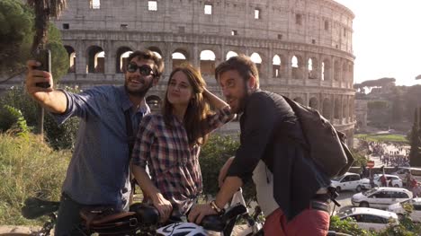 Drei-glückliche-junge-Freunde-Touristen-mit-Fahrrädern-und-Rucksäcke-am-Kolosseum-in-Rom-unter-Selfies-auf-Hügel-bei-Sonnenuntergang-mit-Bäumen-Zeitlupe-steadycam