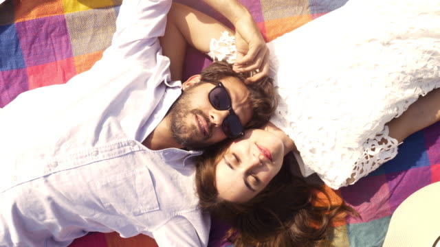 Schöne-junge-Brautpaar-liegend-auf-bunte-Decke-in-den-Park,-Blick-auf-den-Himmel-zeigt-Wolken-romantisch-mit-Gitarre-Sonnenbrillen-schöne-attraktive-Mädchen-Draufsicht-rotierende-Kamera-Slow-Motion