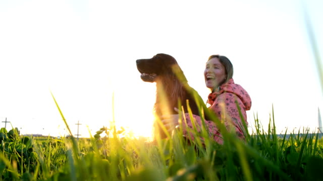 Mujer-abrazando-al-perro-al-atardecer-y-riendo,-niña-con-mascota-sentados-sobre-la-hierba-y-descansar-en-la-naturaleza