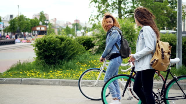 African-American-woman-is-walking-with-her-Caucasian-friend-holding-bicycle-along-beautiful-street-with-fountain-and-wooden-benches,-trees-and-cars-are-visible.