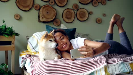 Stolze-Hundebesitzer-glücklich-afroamerikanische-Mädchen-nimmt-Selfie-mit-niedlichen-Haustier-auf-Bett-liegend-in-modernes-Apartment-mit-Smartphone.-Technologie-und-social-Media-Konzept.