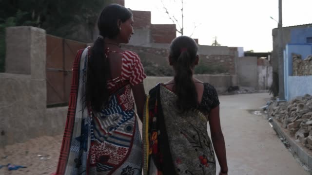 Dos-chicas-a-pie-Paseo-sosteniendo-manos-amigos-bond-calles-casa-rural-ajuste-feliz-compartir-lindo-al-aire-libre-conoce-la-vinculación-siga-cardán-detrás-de-aldea-pequeña-India-vestido-tradicional-traje-ladrillo-muralla