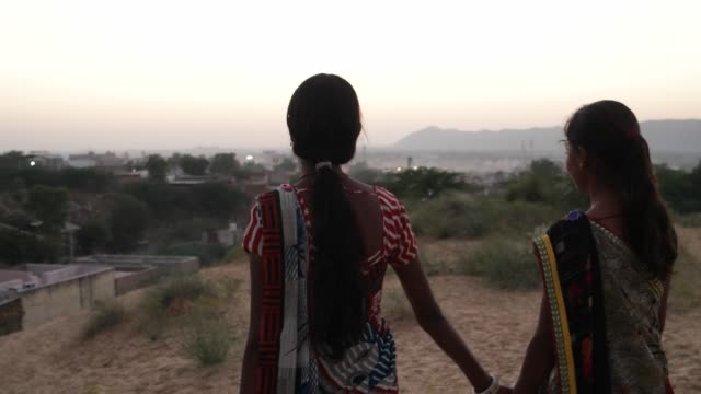 Two-girls-walk-stroll-holding-hands-friends-a-desert-town-in-rural-India-with-sand-and-city-view-evening-dusk-dawn-setting-beautiful-wide-angel-view-handheld-hill-vantage-point-sunset-bonding-follow