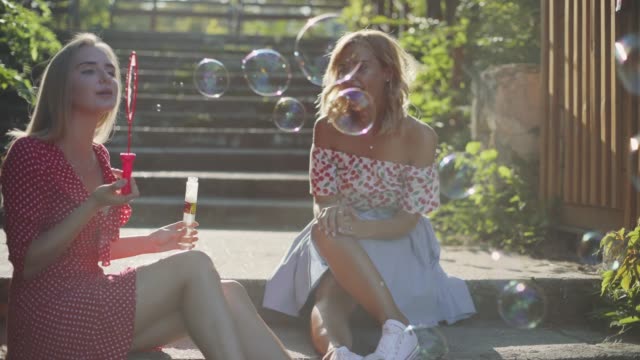 Two-attractive-beautiful-young-blond-women-blow-bubbles-in-the-park-and-are-happy-on-sunset.-Girlfriends-in-park-on-the-steps