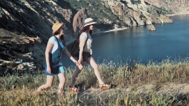 Two-friends-hiker-woman-walking-on-path-holding-hands-enjoying-amazing-mountain-seascape