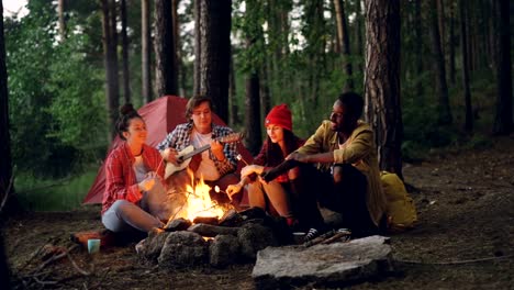 Touristen-ist-das-Gitarrespielen-sitzen-in-der-Nähe-von-Lagerfeuer-mit-Freunden-singen-und-Spaß-haben,-junge-Menschen-sind-Stöcke-mit-Marshmallow-über-Flamme-halten.-Essen,-Musik-und-Spaß-Konzept.