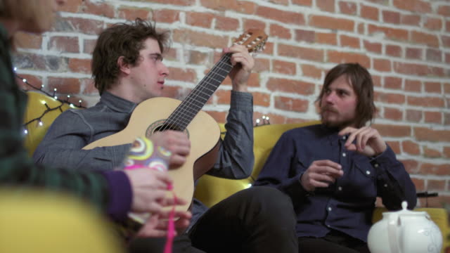 Two-young-men-sitting-on-sofa,-one-playing-guitar