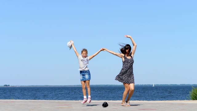 on-beach-mom-and-daughter,-beautiful-brunette-in-sundress-and-girl-child-teenager,-dancing,-listening-to-music-with-mini-music-bluetooth-portable-loudspeaker-on-a-hot-summer-day