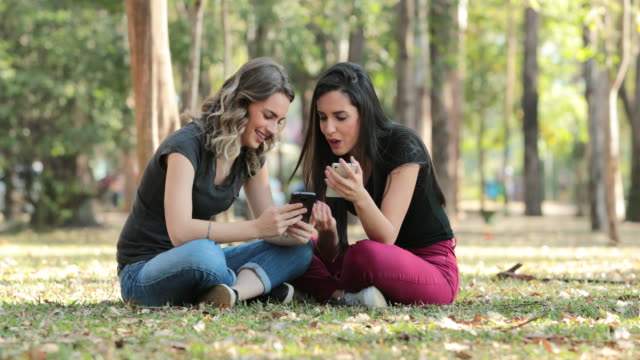 Ehrliche-Freunde-draußen-im-Park-Überprüfung-ihres-Handys.-Mädchen-sitzen-auf-ihren-Smartphones-im-Chat