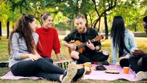Good-looking-guy-is-playing-the-guitar-while-his-male-and-female-friends-are-listening-to-music,-singing-and-having-fun-on-picnic-in-park.-Friendship-and-culture-concept.
