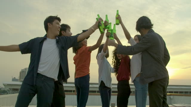 Group-of-young-friends-dancing-and-having-fun-celebrate-with-toast-and-clinking-raising-glasses-at-summer-rooftop-party-with-sunset-back-ground.-Slow-Motion.