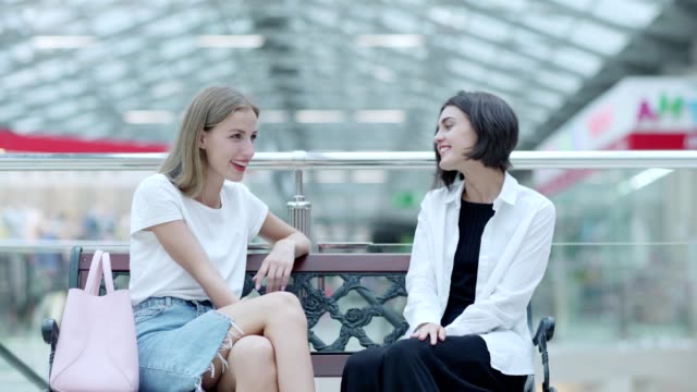 Fade-in-of-two-stylish-girls-resting-on-bench-in-mall-after-shopping,-talking-joyfully-and-laughing