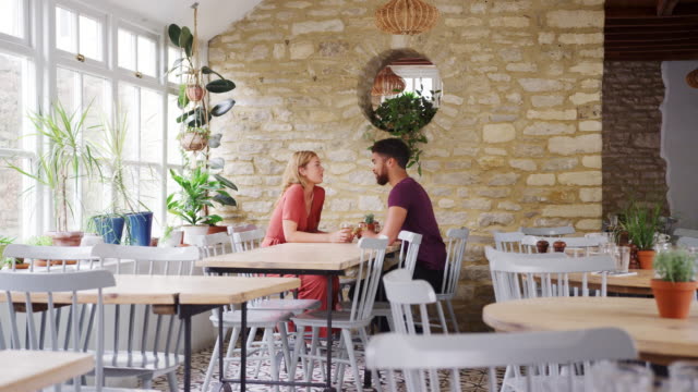 Sonriente-pareja-joven-mestiza-sentados-uno-frente-a-otro-en-una-mesa-en-un-restaurante-vacío