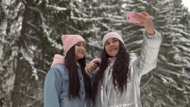 Two-girlfriends-make-selfie-on-the-background-of-the-winter-forest-using-a-smartphone.-4K