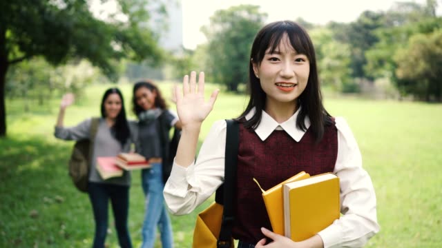 Three-female-women-students-waving-hands-in-the-park-while-focus-is-on-Asian-woman-in-the-front