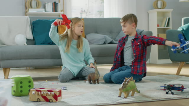 In-the-Living-Room:-Boy-and-Girl-Playing-with-Toy-Airplanes-and-Dinosaurs-while-Sitting-on-a-Carpet.-Sunny-Living-Room-with-Children-Having-Fun.