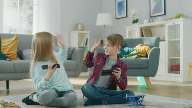 Zu-Hause-auf-einem-Teppich-sitzen:-niedliche-kleine-Mädchen-und-süßen-Boy-spielen-gruppenweise-in-Videospiel-auf-zwei-Smartphones,-hielt-sie-im-horizontalen-Modus-\"Querformat\".-Glückliche-Kinder-tun-High-Five.