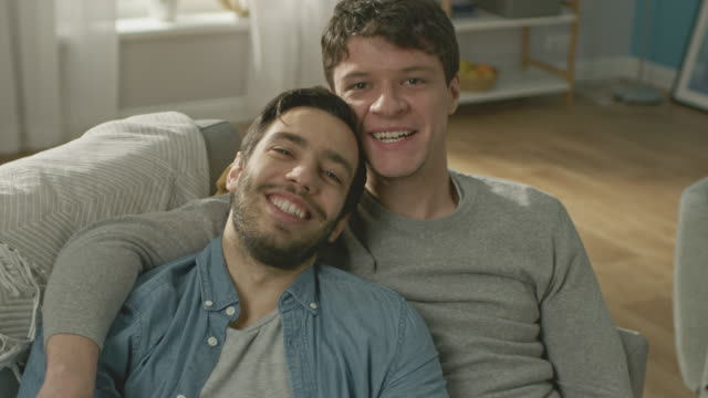 Portrait-of-a-Cute-Male-Queer-Couple-at-Home.-They-Sit-on-a-Sofa-and-Look-at-the-Camera.-Partner-Embraces-His-Lover-from-Behind.-They-are-Happy-and-Smiling.-Room-Has-Modern-Interior.