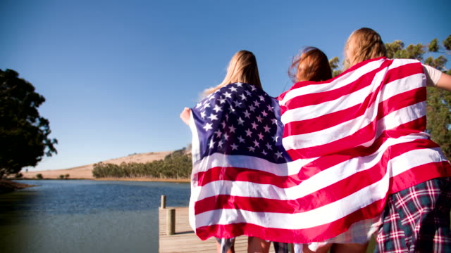 Chicas-adolescentes-corriendo-con-bandera-estadounidens\'en-el-embarcadero-del-lago