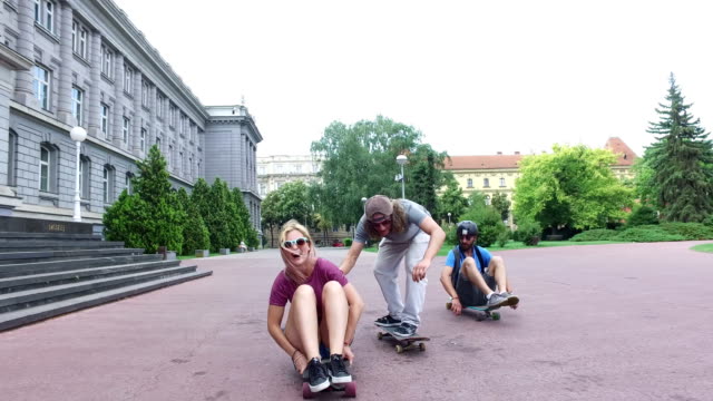 Man-and-woman-sitting-on-a-longboard,-friend-skateboarding-next-to-them