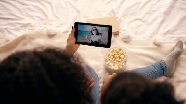 Top-Ansicht-von-zwei-Freundinnen,-die-zu-Hause-auf-dem-Bett-sitzen-und-auf-dem-Skype-auf-dem-Tablet-Computer-mit-ihrem-Freund-Urlaub-am-Strand-sprechen