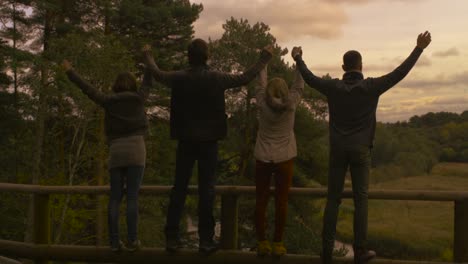 Group-of-people-walk-near-the-edge-of-a-hill,-take-each-other-hands-and-raise-them-while-looking-at-a-forest-landscape-at-sunset.