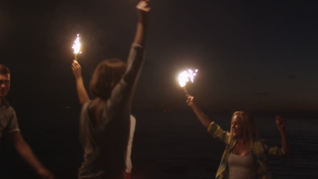 Gruppe-von-Jugendlichen-glücklich-mit-einem-funkelnden-Feuerwerk-in-Händen-laufen-am-Strand