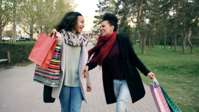 Dolly-shot-of-two-attractive-mixed-race-women-dancing-and-have-fun-while-walking-down-the-park-with-shopping-bags.-Happy-young-friends-walk-after-visiting-mall-sale