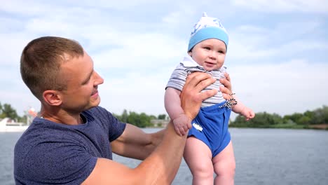 happy-daddy-holds-into-hand-first-child-near-river,-lucky-dad-play-with-baby-on-loch,-little-boy-in-father's-strong-hands