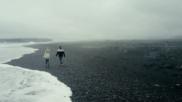 Luftaufnahme-des-jungen-Paares-zu-Fuß-auf-den-schwarzen-Vulkanstrand-in-Island.-Mann-und-Frau-in-Lopapeysa-werfen-Felsen-am-Meer