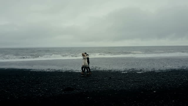 Copter-flying-near-the-young-couple-in-icelandic-sweater.-Man-and-woman-walking,-holding-hands-on-black-volcanic-beach