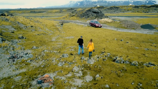 Aerial-back-view-of-young-couple-walking-through-volcanic-lava-field.-Stylish-man-and-woman-traveling-by-car-together