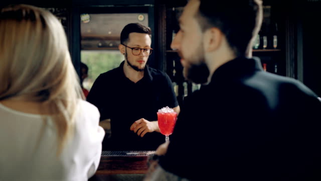 Barman-making-cocktail-for-young-friends-at-the-bar.-A-cheerful-company,-sitting-at-a-bar-with-drinks,-and-talking