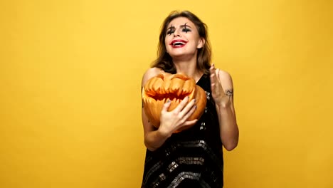 Young-pretty-funny-lady-with-scary-make-up-playing-and-hugging-with-pumpkin-isolated-over-yellow