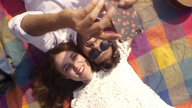 Schöne-junge-Brautpaar-auf-bunte-Decke-im-Park-unter-Selfies-Smartphone-romantisch-mit-Gitarre-Sonnenbrillen-liegen-schöne-attraktive-Mädchen-Draufsicht-rotierende-Kamera-Slow-Motion