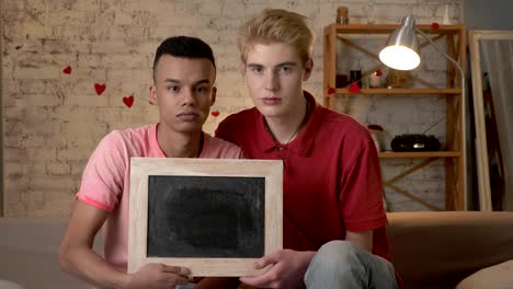 A-sad-international-gay-couple-is-sitting-on-the-couch-and-holding-a-empty-sign.-Home-comfort-on-the-background.-60-fps