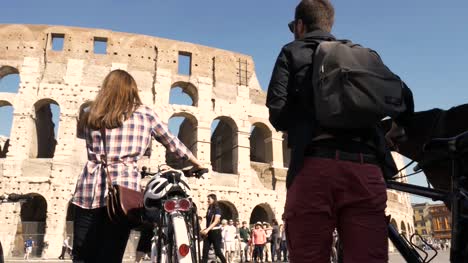 Drei-junge-Freunde-Touristen-zu-Fuß,-mit-Fahrrad-und-Rucksack-im-Colosseum-in-Rom-am-sonnigen-Tag-Zeitlupe-Kamera-Steadycam