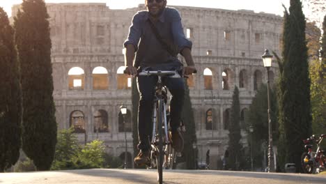 Drei-junge-Freunde-Touristen-Reiten-Fahrräder-in-Colle-Oppio-Park-vor-Kolosseum-auf-Straße-mit-Bäumen-bei-Sonnenuntergang-in-Rom-Slow-motion