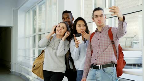 Group-of-multi-ethnic-students-taking-selfie-on-smartphone-camera-while-standing-in-corridor-of-university-.-Hipster-guy-holding-phone-and-friends-are-posing-positively