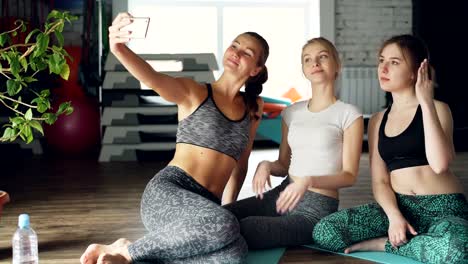 Attractive-young-women-are-taking-selfie-after-yoga-class-in-light-modern-gym-in-wellness-center.-Women-are-wearing-trendy-sports-clothes.-Sports-equipment-in-background.