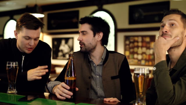 Cheerful-handsome-guys-are-having-conversation-in-bar-over-beer.-Young-men-are-holding-bottles-and-glasses,-chatting-and-gesturing-emotionally.