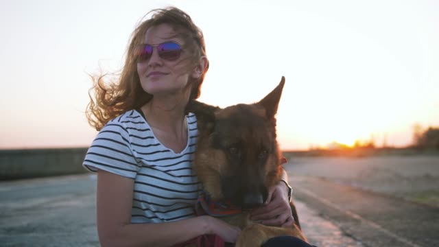Glückliche-junge-Frau-halten-und-umarmen-ihr-deutscher-Schäferhund-im-Freien-auf-dem-Meer-Pier-bei-Sonnenuntergang