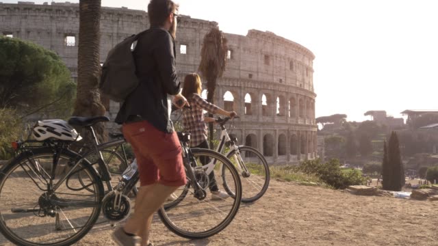 Drei-glückliche-junge-Freunde-Touristen-mit-Fahrrädern-und-Rucksäcke-am-Kolosseum-in-Rom-ankommen-auf-Hügel-bei-Sonnenuntergang-mit-Bäumen-Zeitlupe-steadycam