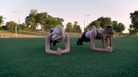 Two-young-beautiful-womans-doing-plank-exercise-on-soccer-field-in-park-during-morning,-fitness-models-exercising